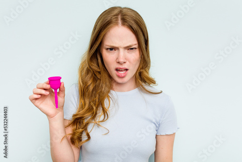 Young caucasian woman holding menstrual cup isolated on blue background screaming very angry and aggressive.