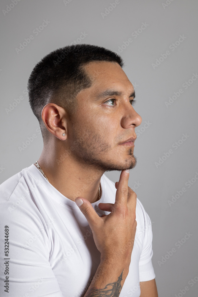 face of a young latin man with short urban style hair with a beard and a white t-shirt in studio has his index finger on his chin, thinking face of a young latin man with short urban style hair with a beard and a white t-shirt in studio has his index finger on his chin, thinking