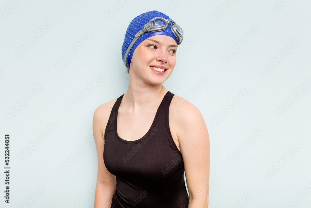 Young caucasian swimmer woman isolated on blue background looks aside smiling, cheerful and pleasant. Young caucasian swimmer woman isolated on blue background looks aside smiling, cheerful and pleasant.