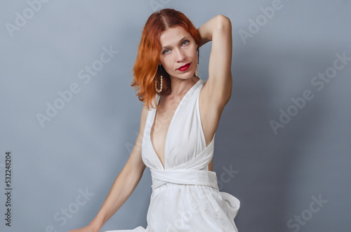 Portrait of a red-haired woman in a stylish off-shoulder white dress. Looks into the camera. Hand is raised behind the head.