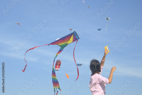 A little girl is playing in the wind with a kite. Other kites are in the blue sky.