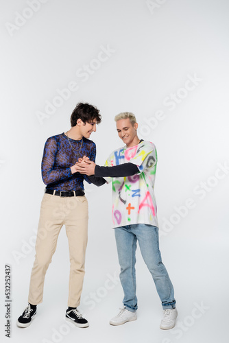 full length of positive gay holding hands of stylish queer person on grey background.
