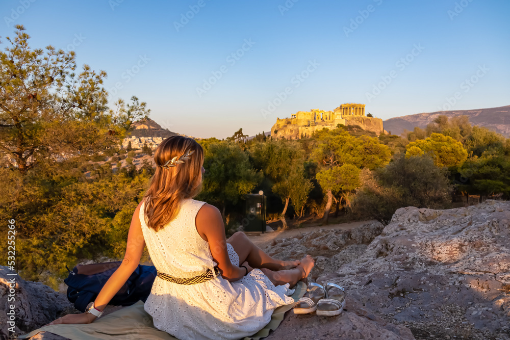 Rear view of tourist woman wearing a white dress and golden laurel crown looking at Parthenon of the Acropolis of Athens during sunset seen from Pnyx Hill, Attica, Greece, Europe. Ruins ancient temple