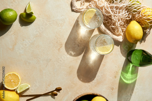 Fresh citrus in mesh bag and glasses of lemon water on beige background. Lemons and limes in string shopping bag with tonic water, top view, copy space photo