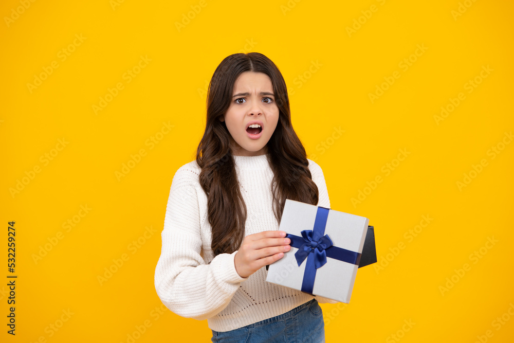 Teenager child holding gift box on yellow isolated background. Gift for kids birthday. Christmas or New Year present box. Angry face, upset emotions of teenager girl. Teenager child holding gift box on yellow isolated background. Gift for kids birthday. Christmas or New Year present box. Angry face, upset emotions of teenager girl.