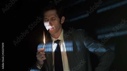 A serious young man lights his pipe with a match, a man in a gray suit with a tie holds a burning match in his hand, deep shadows in a retro noir style, light through the blinds photo