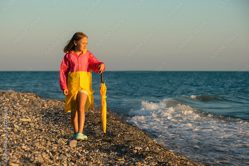 a schoolgirl dressed in a bright colored raincoat and with a yellow umbrella walks along the shore of a bright blue sea and looks at the waves