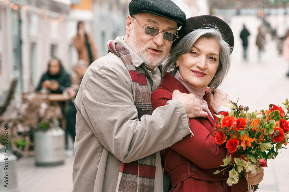 Obraz premium Elderly senior love couple. Old retired man woman together on romantic date.Aged husband wife walking on city street with flowers.Stylish elder hugging people pensioner in red coat.Happy family years