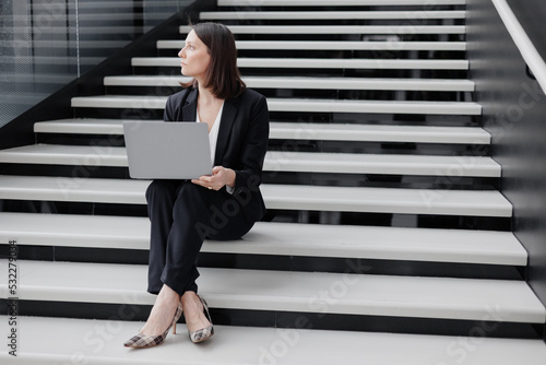 business woman in a formal suit with a laptop in an office or business center sits on the steps in the lobby and works online. Work in a corporation as a marketing financier or lawyer. a successful