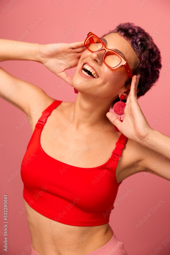 Young athletic woman with a short haircut and purple hair in a red top and pink yoga leggings in sunglasses with an athletic figure smiles and dances on a pink background Young athletic woman with a short haircut and purple hair in a red top and pink yoga leggings in sunglasses with an athletic figure smiles and dances on a pink background