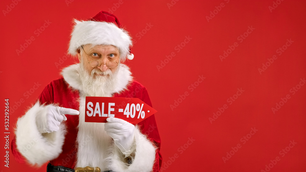 Santa Claus Holds Sign With Inscription Sale -40 off, Points His Finger at an Empty Space Mock up and Looks at Discount in Camera and Smiles on Red Background. Big Discount, Christmas Holidays Sales.