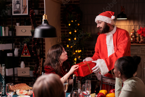 Man wearing santa claus costume giving woman christmas gift  happy wife getting present giftbox from husband. Xmas night celebration with big family at festive dinner table  winter holidays greeting