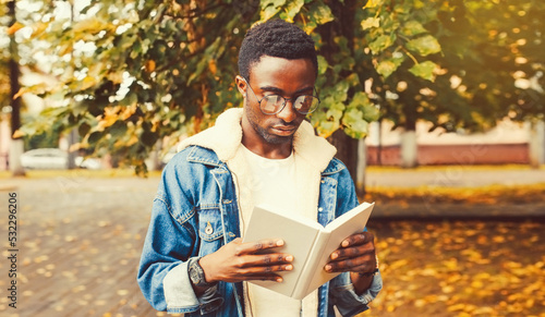 Portrait of young african man student reading a book wearing eyeglasses in autumn city park