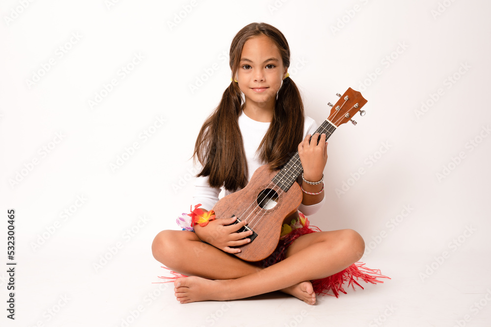 Cute little girl in summer clothing playing ukulele sitting on the floor  over white background. Happy vacation concept. Stock Photo