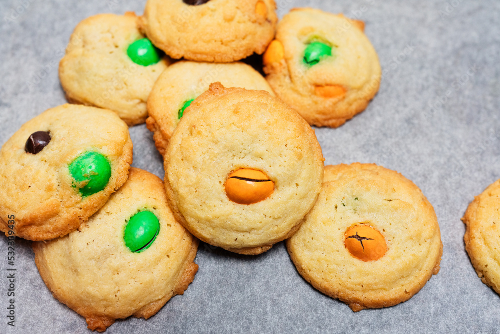 Typical American sugar cookies with chocolate covered candies in Halloween treats colors orange and green on baking sheet, easy and fun biscuit baking recipe for kids for trick or treating sweets