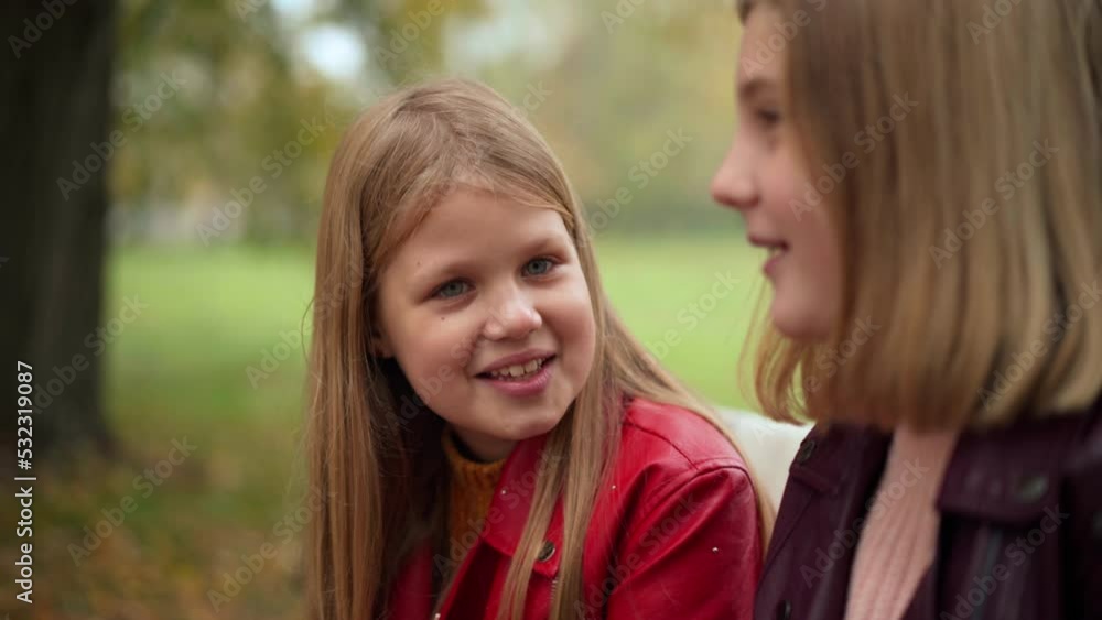 Curios positive Caucasian teen girl with toothy smile talking to friend sitting on bench in autumn park. Portrait of happy confident relaxed teenager sharing gossips outdoors looking away smiling