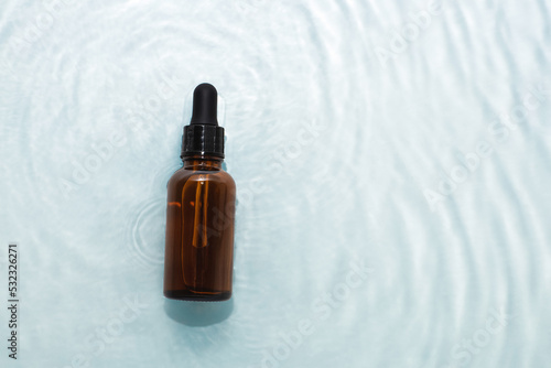 Dark bottle of serum with dropper lies in water with waves on the surface, copy space