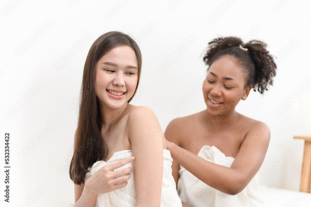 Young diversity ethnic beautiful woman in white towel after shower bath help each other apply body skin care lotion moisturizer in bedroom. Woman daily lifestyle or LGBTQ couple love concept. Young diversity ethnic beautiful woman in white towel after shower bath help each other apply body skin care lotion moisturizer in bedroom. Woman daily lifestyle or LGBTQ couple love concept.