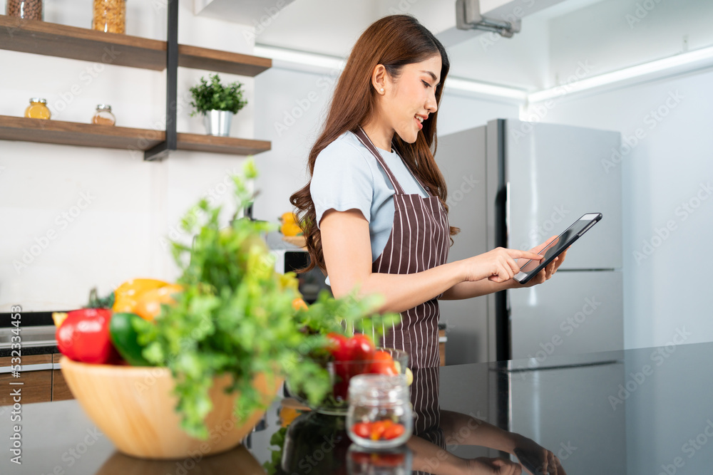 Happy Asian woman wearing an apron prepares vegetables for healthy food by searching recipes online on a digital tablet in home kitchen, watching videos of ingredients. Modern cooking technology.