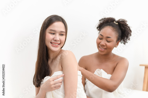 Young diversity ethnic beautiful woman in white towel after shower bath help each other apply body skin care lotion moisturizer in bedroom. Woman daily lifestyle or LGBTQ couple love concept.