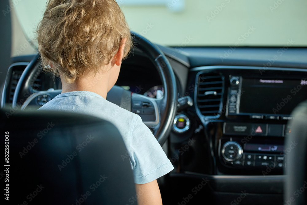 View from behind curly boy sitting behind the wheel of car in driver's seat and looking forward into the windshield. Child dreams of becoming a racer, sitting behind the wheel of his father's car