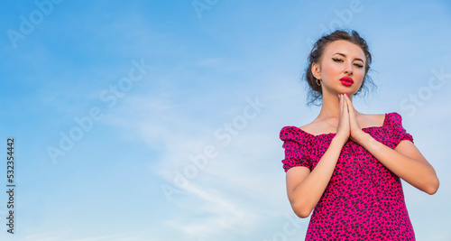 Young pretty and tanned girl in burgundy dress stands against background of blue cloudy summer sky, hands clasped together under her chin. Looks at camera.