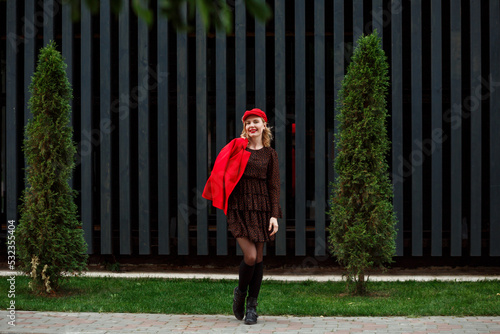 Full length portrait of fashionable woman in red cap, red jacket, dark dress, black boots with gaiters in front of modern wooden building outdoors in the city streets. 