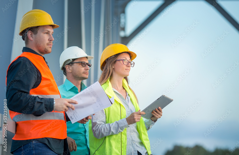 Railway and road engineers are standing in orange and white helmets and signal vests with documents against the background of the metal structure of the bridge. Railway and road engineers are standing in orange and white helmets and signal vests with documents against the background of the metal structure of the bridge.