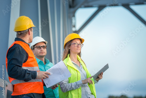 young men and a woman are standing in orange and white helmets and signal vests with documents against the background of the metal structure of the bridge. The concept of railway and road engineers..