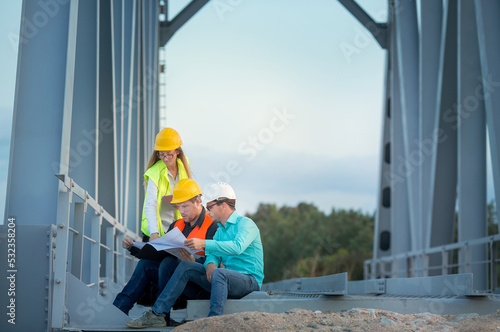 young men and a woman are standing in orange and white helmets and signal vests with documents against the background of the metal structure of the bridge. The concept of railway and road engineers..
