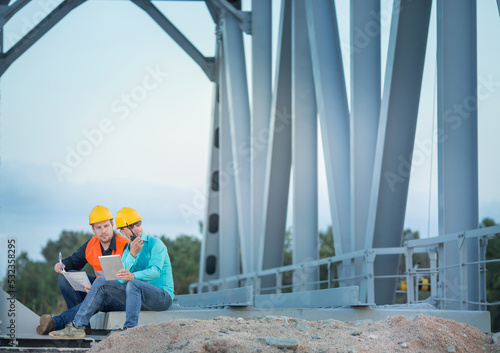 young men  are sitting  in orange and white helmets and signal vests they are sitting with documents and a tablet on a metal bridge, , talking into a walkie-talkie.    photo