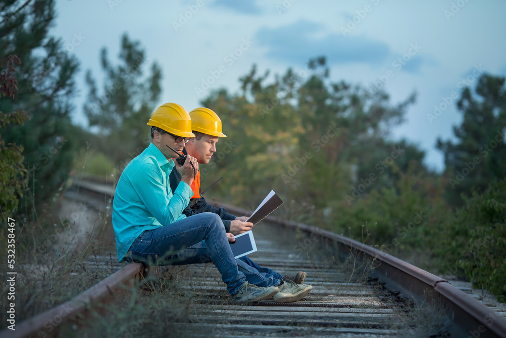 Two young men with documents, walkie-talkies and yellow helmets are sitting on the rails of the railway