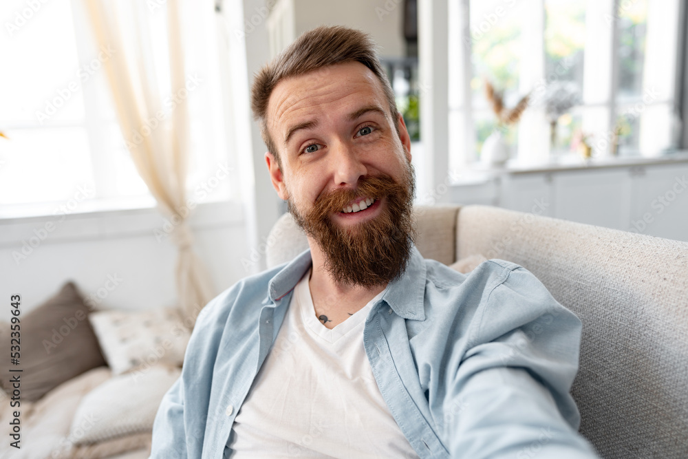Bearded happy young man smiling and taking selfie photo while sitting on sofa at home. Bearded happy young man smiling and taking selfie photo while sitting on sofa at home.