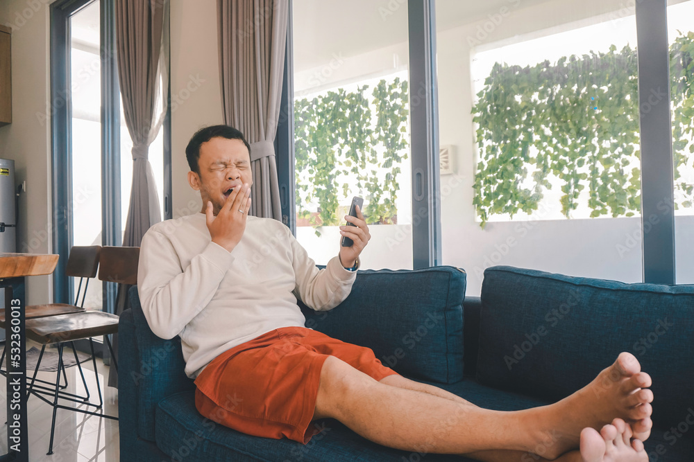Obraz premium A young Asian man spends his free time at home sitting on a cozy sofa in the living room yawning while holding his smartphone