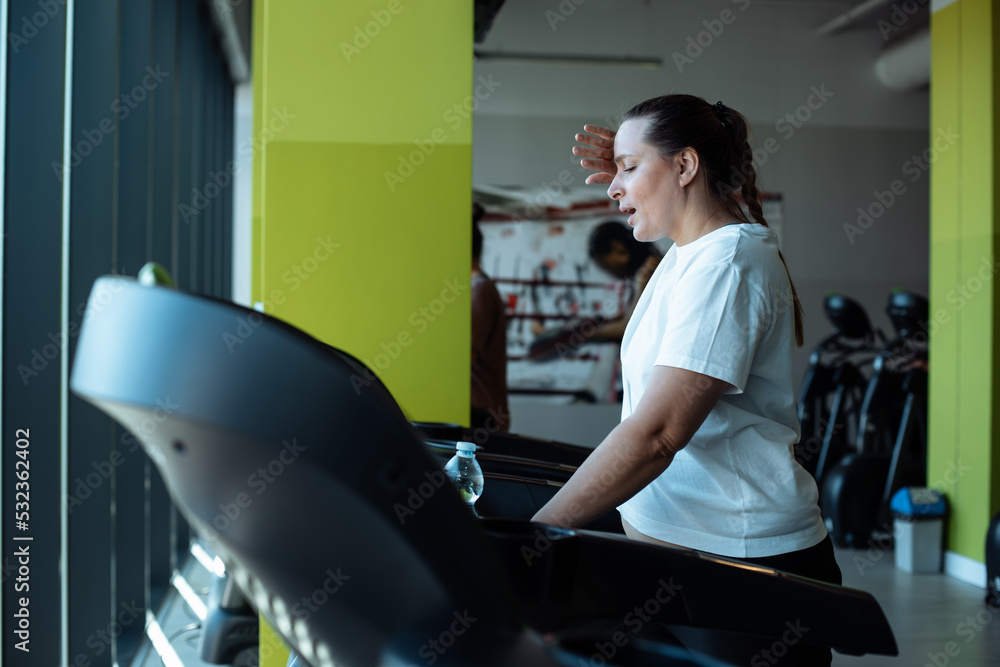 Tired plus size woman run on treadmill at fitness gym wiping sweat from face by hand, side view. Middle aged lady do cardio workout on exercise machine. Weight loss, gym membership, sport lifestyle Tired plus size woman run on treadmill at fitness gym wiping sweat from face by hand, side view. Middle aged lady do cardio workout on exercise machine. Weight loss, gym membership, sport lifestyle
