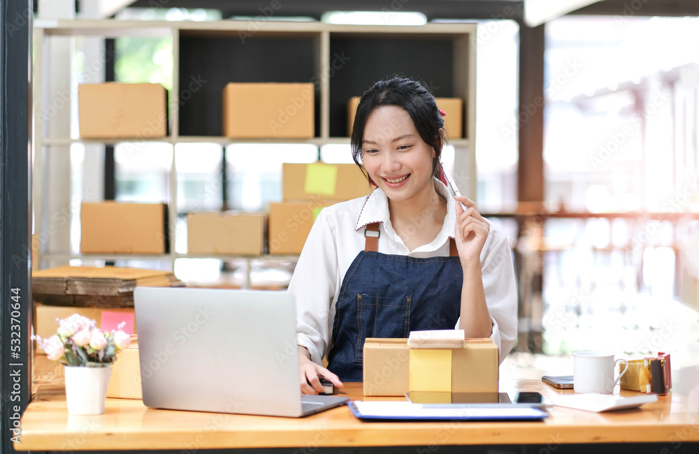 Startup SME small business entrepreneur of freelance Asian woman wearing apron using laptop and box to receive and review orders online to prepare to pack sell to customers, online sme business ideas. Startup SME small business entrepreneur of freelance Asian woman wearing apron using laptop and box to receive and review orders online to prepare to pack sell to customers, online sme business ideas.