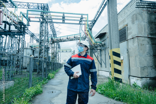 A masked power engineer during a pandemic inspects the modern equipment of an electrical substation before commissioning. Energy and industry. Scheduled repair of electrical equipment.