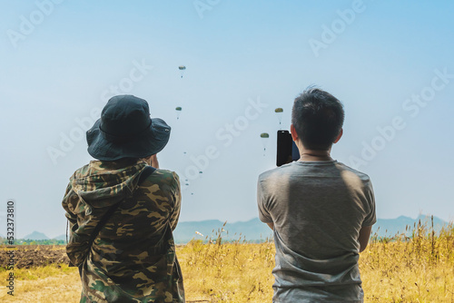 Parents take video clips and photo with smart phone and watch with worry and concern during parachute training from airplane for army cadet with blurred image of parachute and landscape in background.