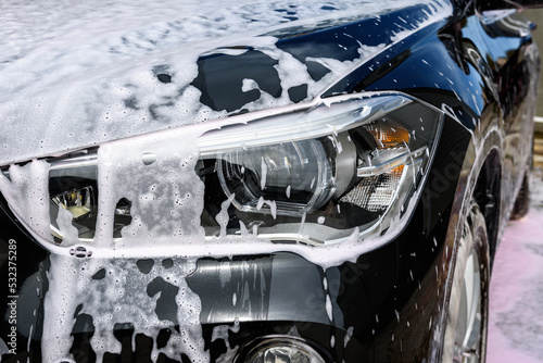 Black car in white soapy foam outdoor. Vehicle cleaning with detergent.