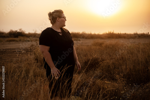 Beautiful overweight woman of xl size dressed in a black dress posing in a field with dry grass against the background of a magnifivent sunset © Вероника Зеленина