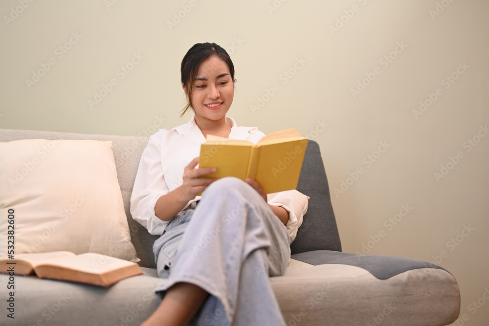 Pleasant millennial woman reading a book, relaxing on couch in bright living room Pleasant millennial woman reading a book, relaxing on couch in bright living room