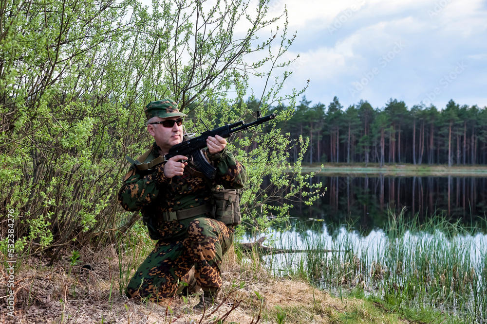 Russian soldier man dressed military camouflage uniform with weapon in forest with lake at nature background. Male border guard in country border with autogun on war. Copy text space