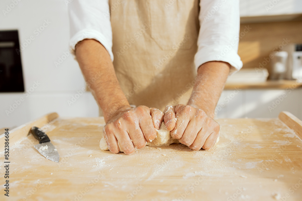 Close-up shot of men's hands kneading dough on wooden surface preparing homemade bread pasta pizza in modern light kitchen.