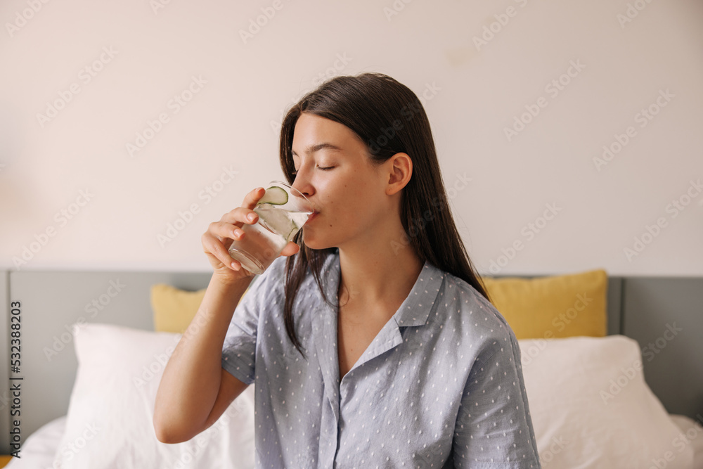 Pretty young caucasian woman drinking water with slice of cucumber sit on bed good morning. Brunette hair girl wears in grey pajamas. Concept healthy lifestyle Pretty young caucasian woman drinking water with slice of cucumber sit on bed good morning. Brunette hair girl wears in grey pajamas. Concept healthy lifestyle