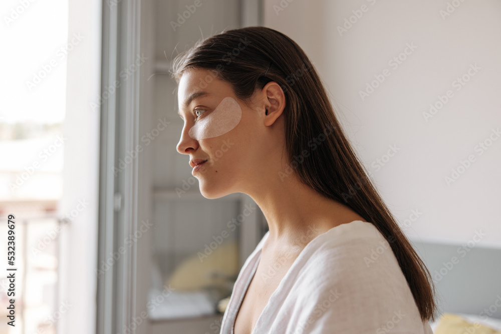 Close-up young caucasian woman with white eyes patches sitting on bed. Brunette hair girl wears in white pajamas looking at distance at home. Concept morning routine.
