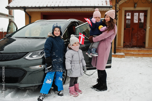 Young dannish mother with kids hold Denmark flags and charging electric car in the yard of her house at winter. photo