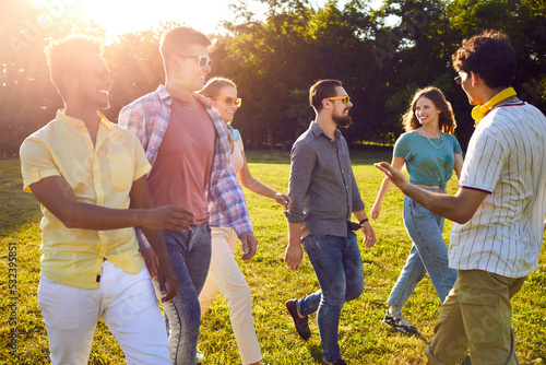 Group of cheerful friends hanging out in a beautiful park. Six happy young diverse multiethnic people enjoying good summer weather, walking on a green lawn, talking and having fun together