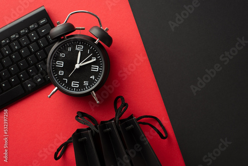 Cyber monday concept. Top view photo of black alarm clock keyboard and paper bags on bicolor red and black background with empty space