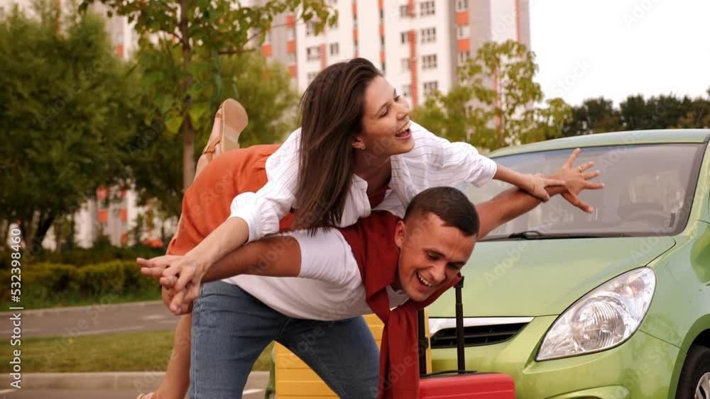 Close-up of a young couple having fun in the parking lot near the car against the background of a multi-storey building, the girl is lying on the guy's back and has her arms out to the sides. Close-up of a young couple having fun in the parking lot near the car against the background of a multi-storey building, the girl is lying on the guy's back and has her arms out to the sides.