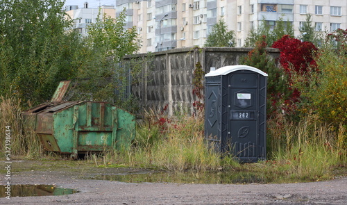 Mobile plastic toilet and dumpster near concrete fence, Bolshevikov Avenue, St. Petersburg, Russia, September 2022 photo